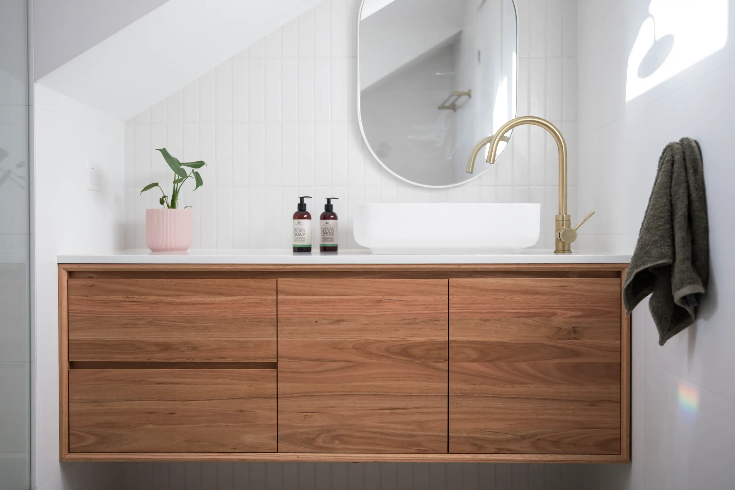 Floating wooden bathroom vanity with white vessel sink, gold faucet, oval mirror, potted plant and towel - Bathroom Cabinets Installation