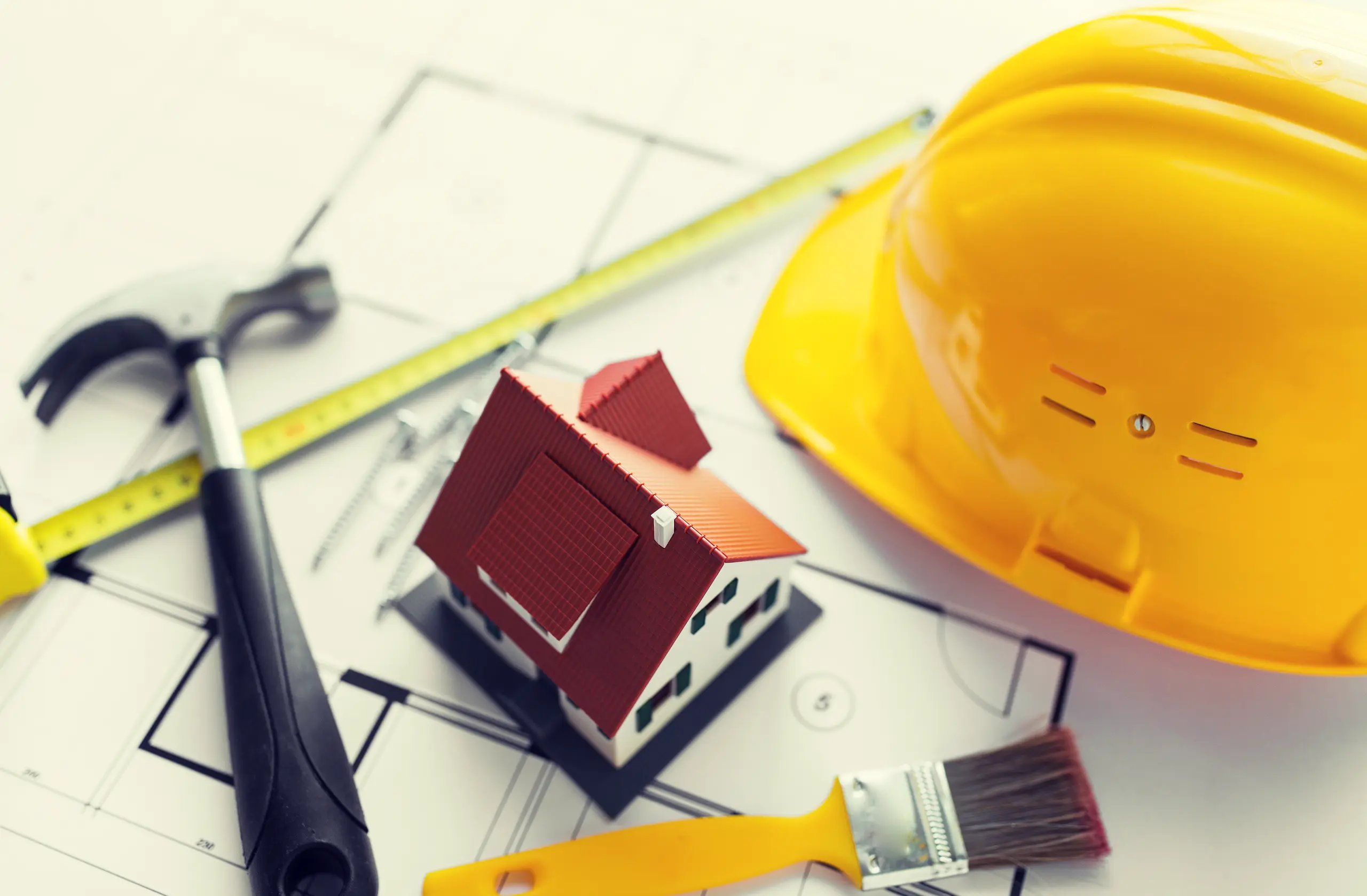 Mini house model on blueprint with hammer, measuring tape, paintbrush and hard hat illustrating Home Remodeling services