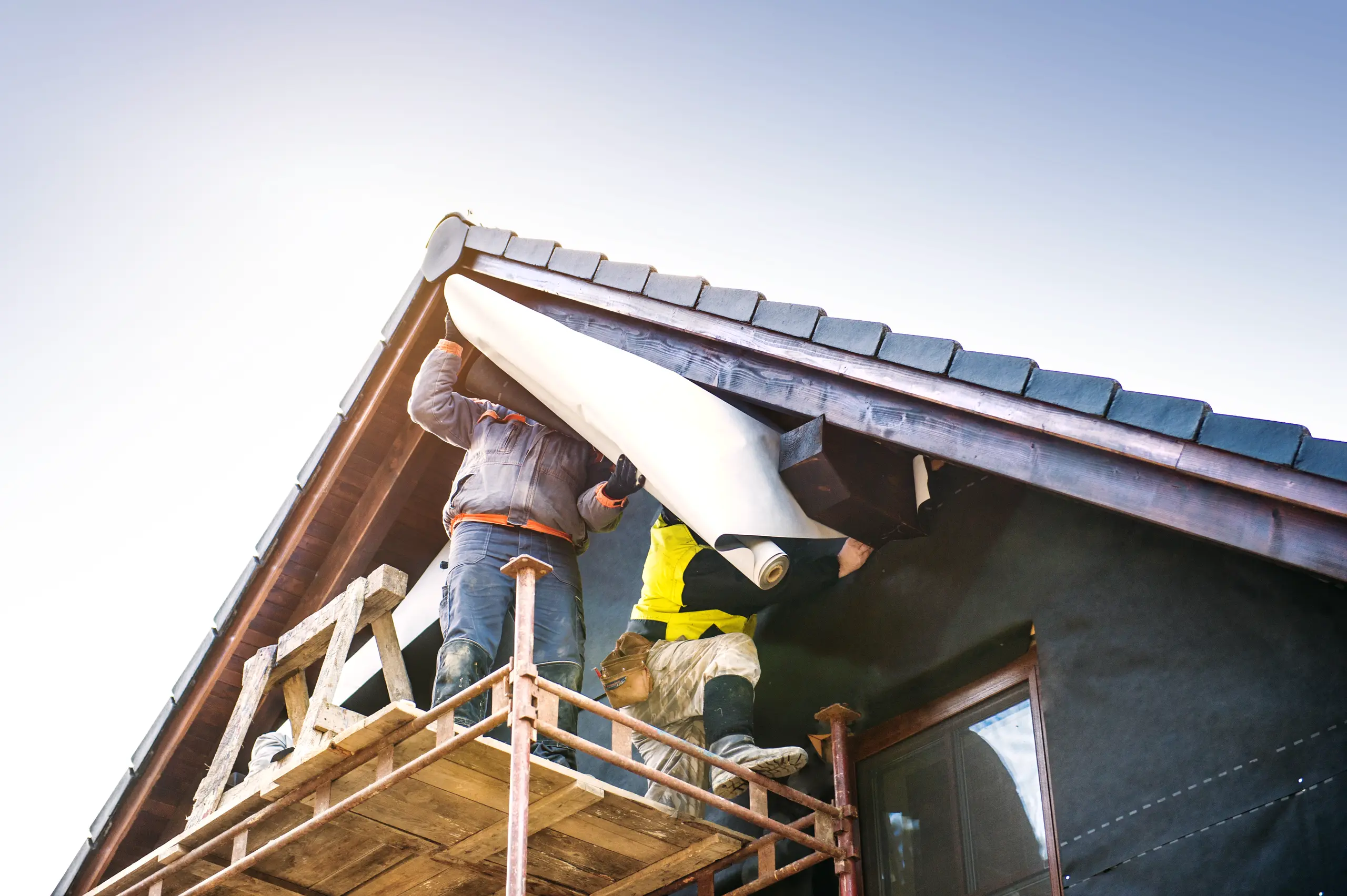 Two workers on scaffolding installing roof underlayment along eaves during exterior home renovation