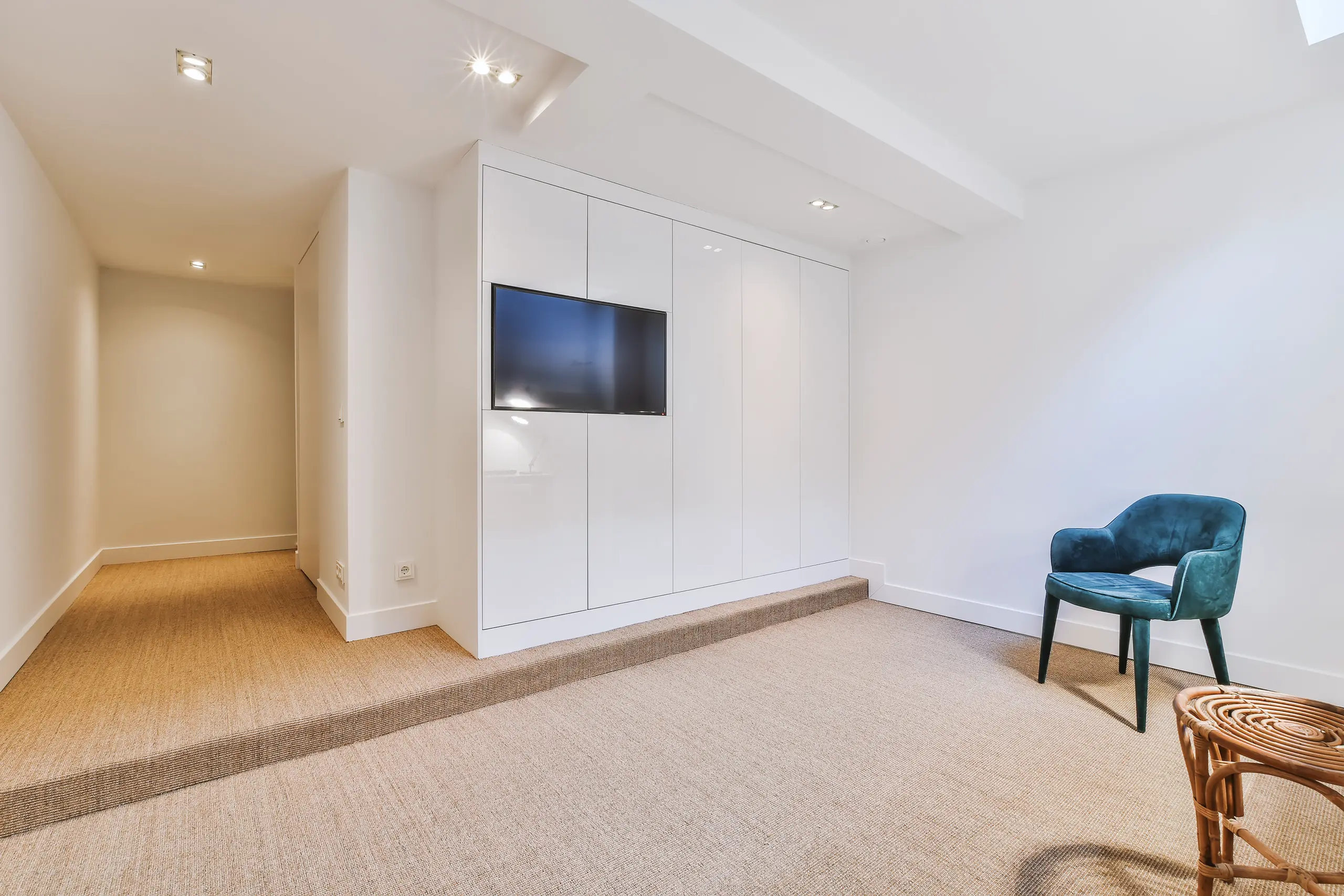 Bright finished basement with white built-in cabinets, wall-mounted TV, beige carpet and teal chair, Basement Finishing