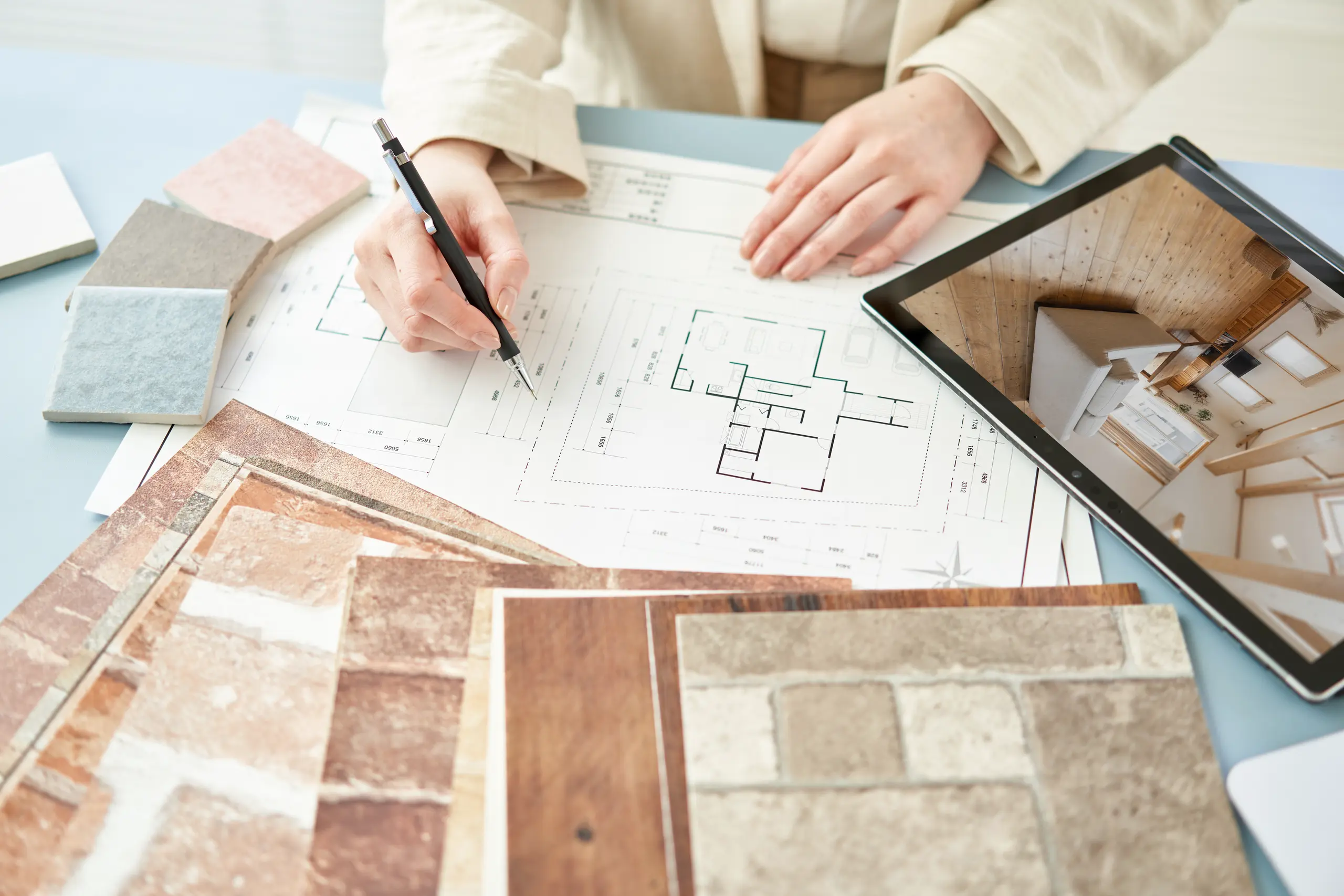 Hand holding pencil over home floor plans with tile samples and tablet, illustrating Tile Work planning