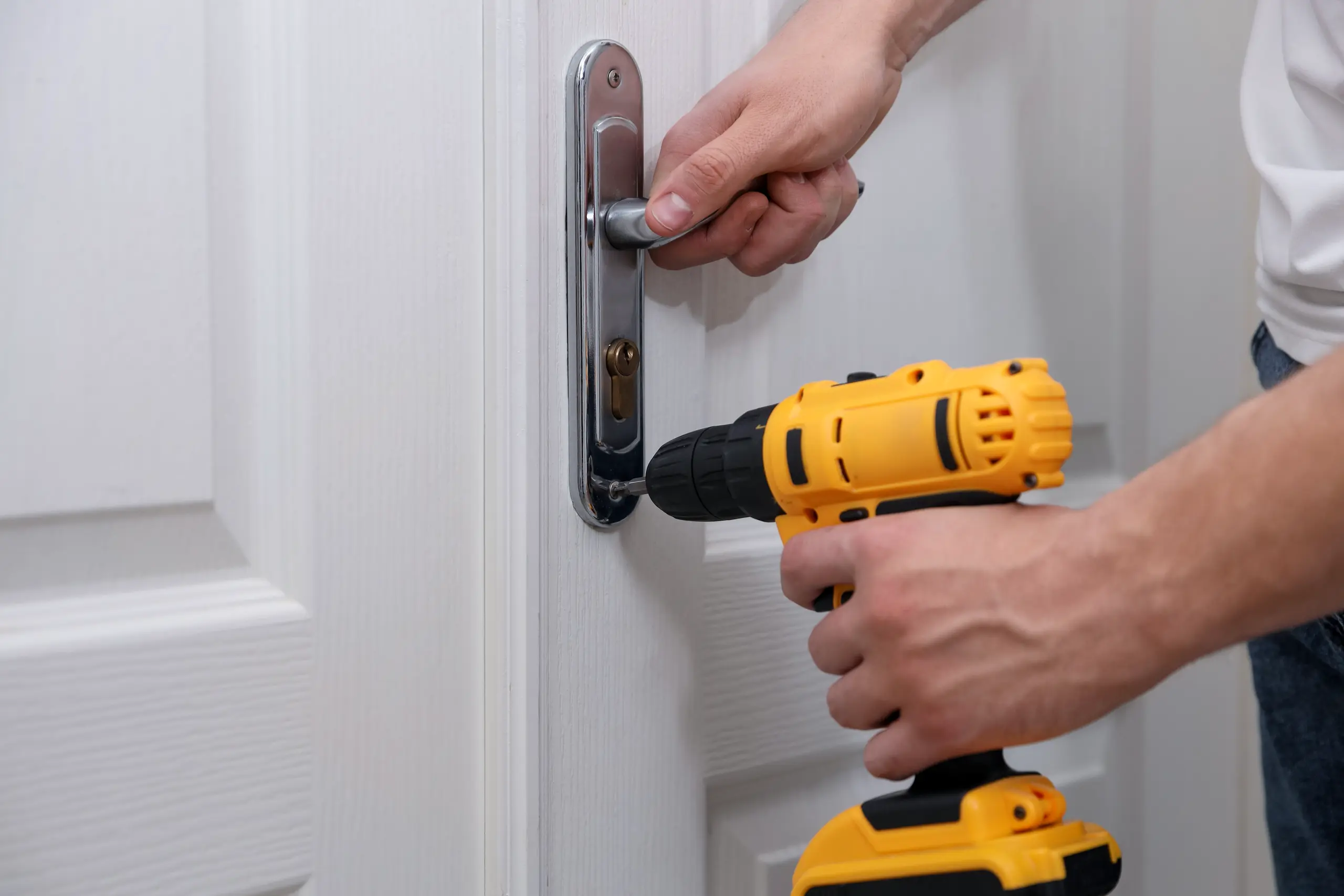 Worker using cordless drill to install chrome door handle and lock on white interior door, Door Installation