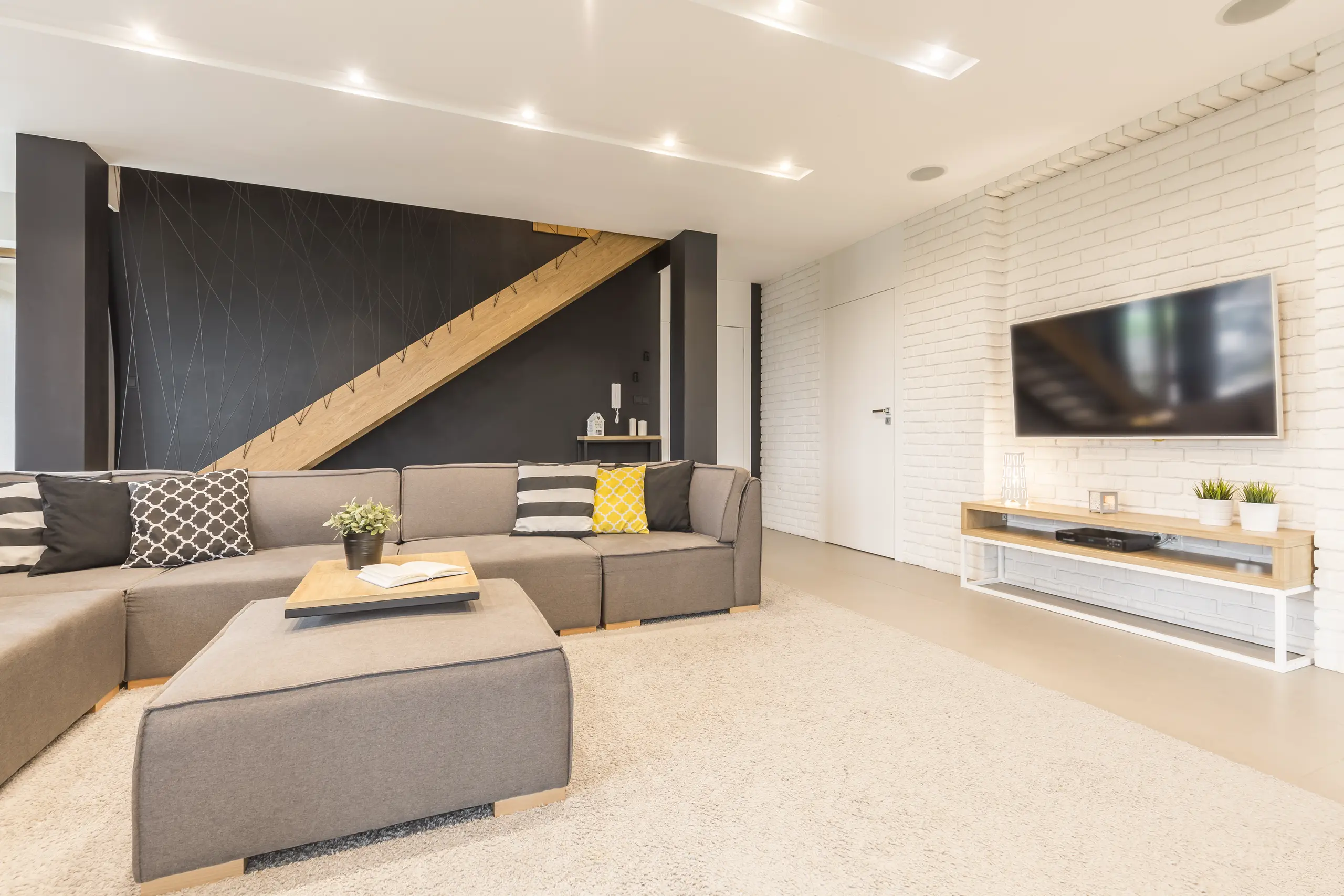 Modern living room after home remodeling with gray sectional sofa, decorative pillows, recessed lighting, staircase and TV wall