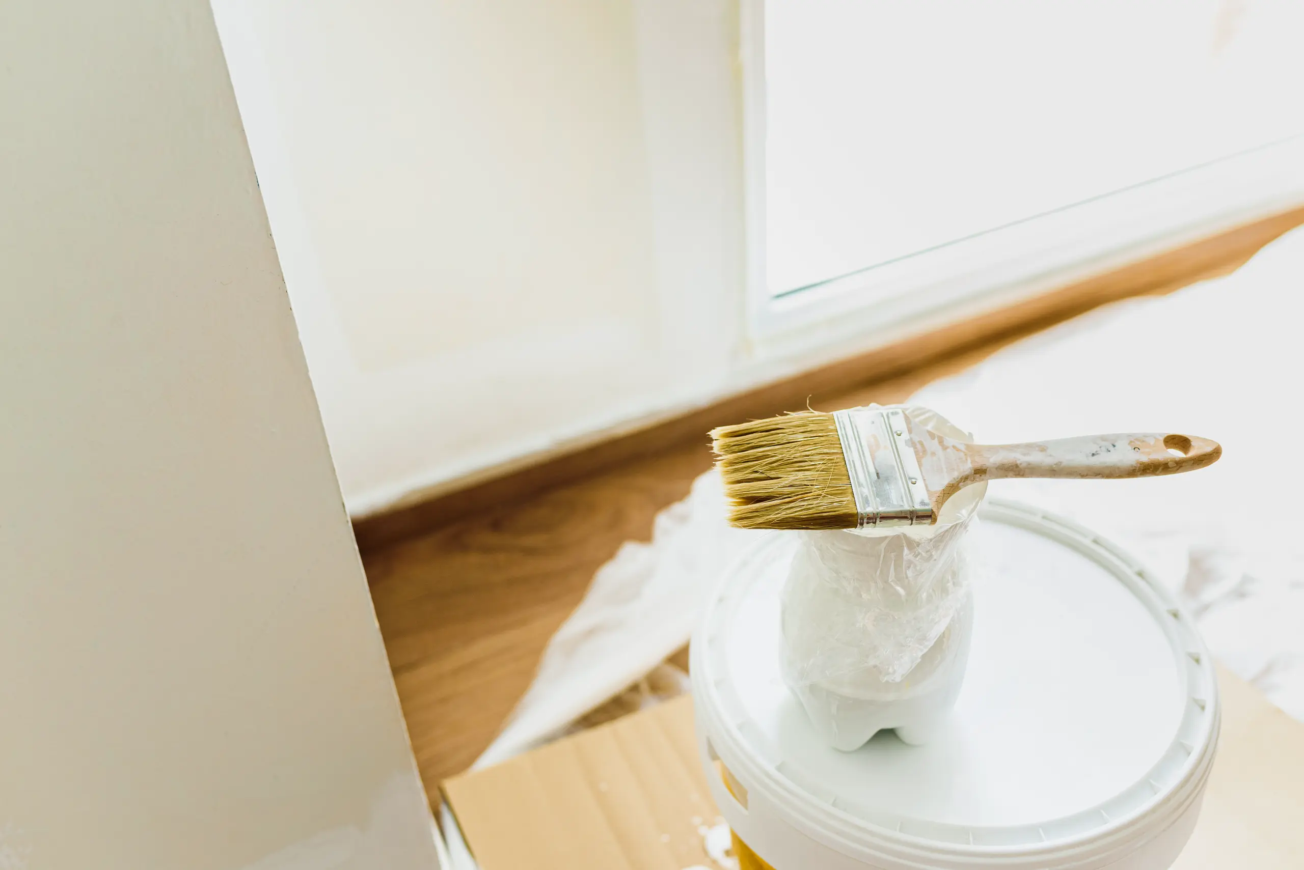 Paintbrush on paint-filled bucket with drop cloth near window for interior painting service