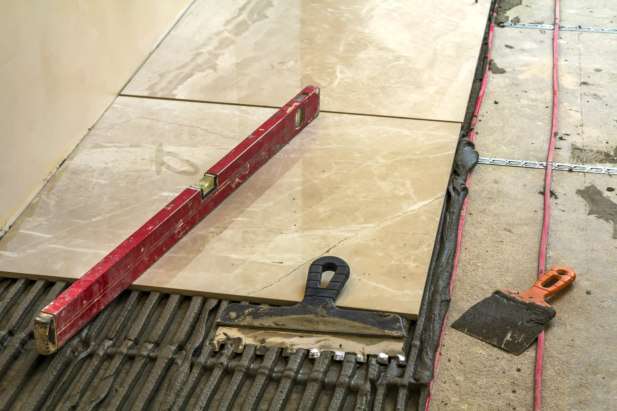 Red spirit level and trowel laying beige floor tiles on mortar bed during Tile Work