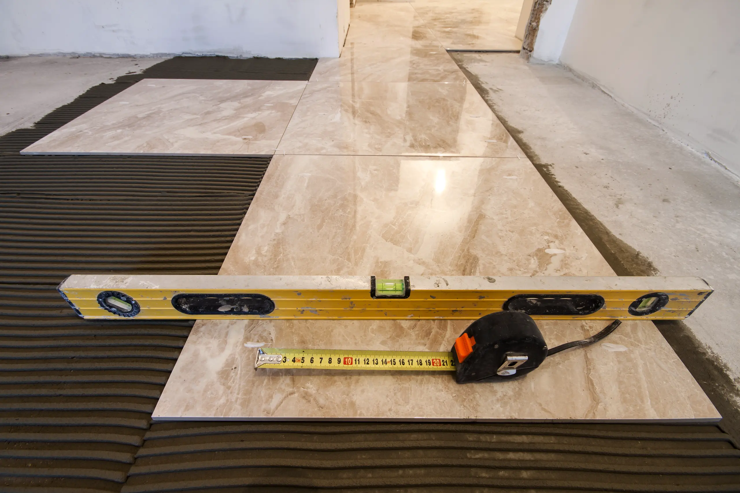Tile Setting large marble-effect floor tiles on adhesive mortar with ridges, featuring a spirit level and tape measure
